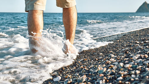 Male legs walking on pebble beach along the shore near the water with waves, low section.