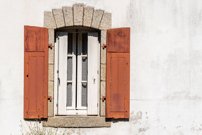 Old window with wooden red painted shutters on white wall with stone jambs. the shutters are open