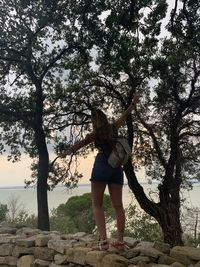 Rear view of woman with arms outstretched standing on retaining wall against trees