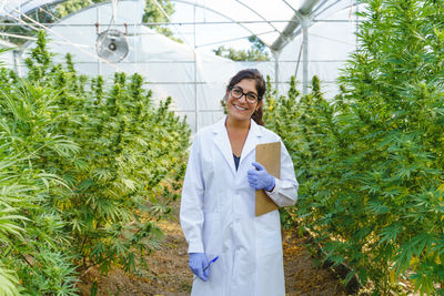 Portrait of smiling young professional female scientist in white robe and gloves standing among bushes of hemp plants in glasshouse in pharmaceutical laboratory and looking at camera