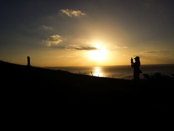 Silhouette woman photographing at beach against sky during sunset