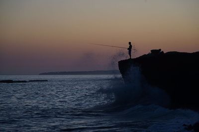 Silhouette person fishing while standing on rock at beach against sky during sunset