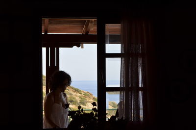 Woman looking at potted plant while standing by window at home