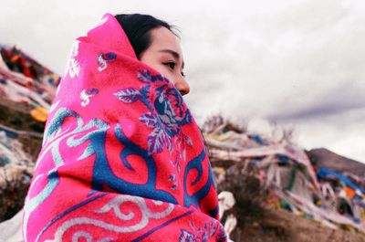 Side view of young woman wrapped in shawl against cloudy sky