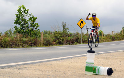 Bottles against male cyclist gesturing while cycling on road against sky