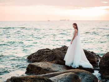 Full length of bride standing on rock in sea against sky during sunset