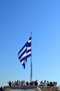 Low angle view of people standing by greek flag against clear blue sky