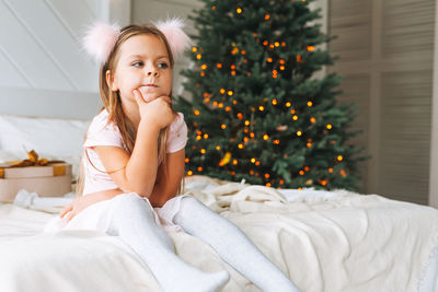 Cute sad little girl in pink dress with present gift box sitting on bed in room with christmas tree