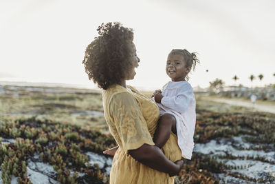 Pregnant mother holding young toddler girl on belly at beachg