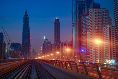 Illuminated railroad tracks amidst buildings in city against sky