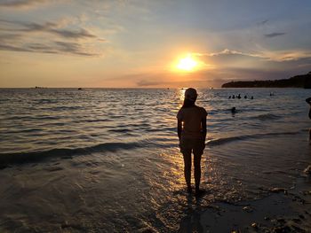 Rear view of woman standing on shore at beach against sky during sunset