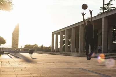 Young man playing basketball on footpath against clear sky at sunset