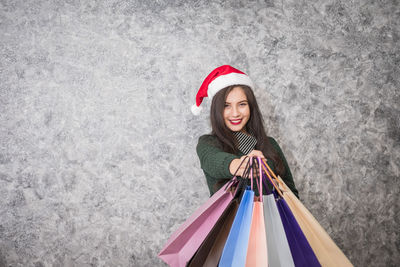 Portrait of woman in santa hat holding shopping bag while standing against wall