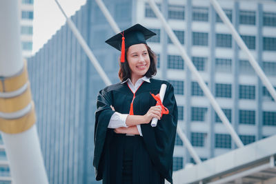 Young woman wearing graduation gown while standing against office building