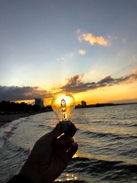 Cropped hand holding light bulb against sea during sunset