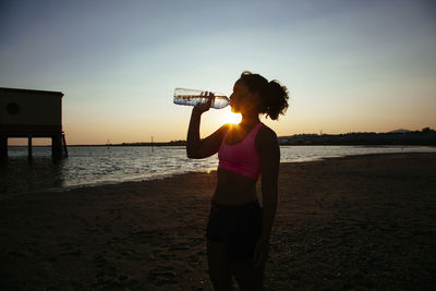 Woman standing at beach and drinking water against sky during sunset