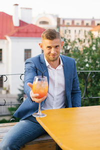 Young handsome man is sitting in an outdoor summer cafe in a blue suit and holding a cocktail 