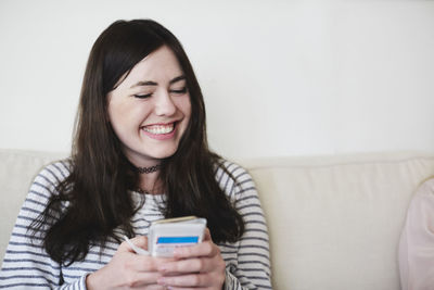 Smiling young woman using mobile phone while sitting on sofa against wall at home