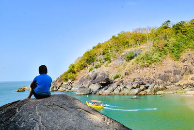 Rear view of boy sitting on rock against blue sky