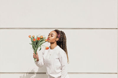 Young woman with eyes closed smelling flowers in front of white wall during sunny day