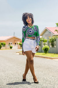 Full length portrait of happy beautiful woman walking on road by house against sky