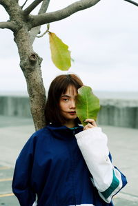 Portrait of woman with leaf standing by tree on footpath