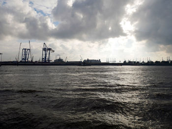 Distant view of cranes at harbor against cloudy sky