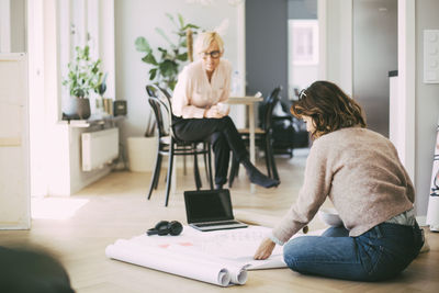 Woman sitting on the floor discussing blueprint with female coworker at home