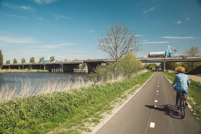 Rear view of woman riding bicycle on road by river against sky