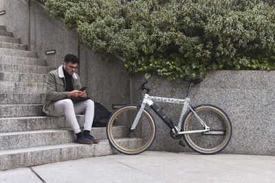 Young man with bicycle using smart phone while sitting on steps