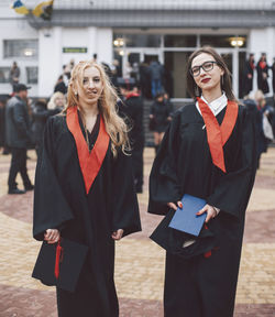 Portrait of happy female students wearing graduation gowns while standing against building