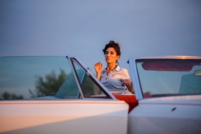 Portrait of young woman sitting on car against sky