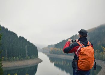 Rear view of man photographing at river against sky