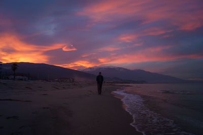 Man walking at beach against cloudy sky during sunset