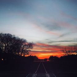 Silhouette trees by road against sky during sunset