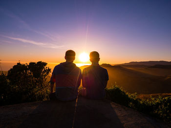 Rear view of silhouette friends sitting on mountain against sky during sunset