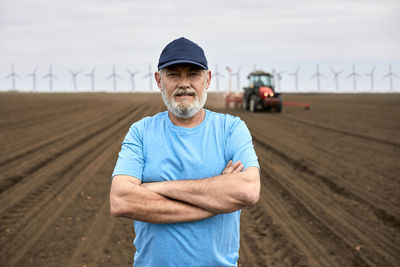 Bearded male farmer with arms crossed standing on plowed agricultural field