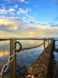 Metal railing by sea and ocean pool against sky with reflections during sunset