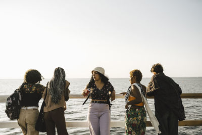 Teenage girl standing amidst multiracial friends leaning on railing against clear sky