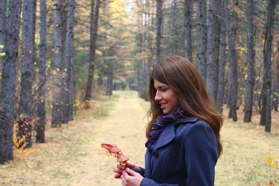 Side view of young woman holding leaf while standing in forest