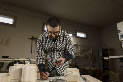 Carpenter holding smart phone writing in note pad at workshop