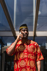 Serious adult black man in traditional red clothes and kufi standing near building with glass walls while having conversation on phone in sunny day in city street and looking away