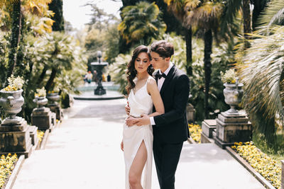 Happy lovers the bride and groom in wedding outfits walk among plants and palm trees in the old park