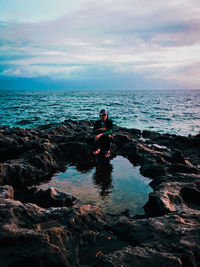 Woman sitting on rock at sea shore against sky