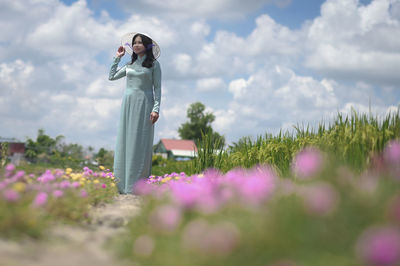 Woman standing by flowering plants on field against cloudy sky