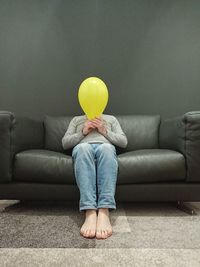 Full length of man holding balloon over face while sitting on sofa at home