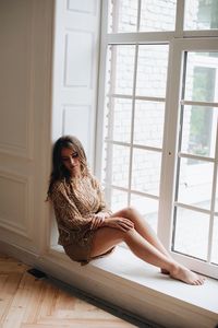 Full length of thoughtful woman sitting on window sill at home