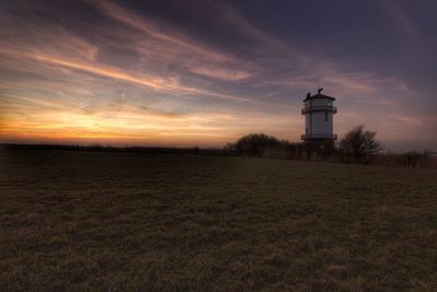 Lighthouse on field by building against sky during sunset