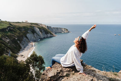 Woman sitting on cliff by sea against sky