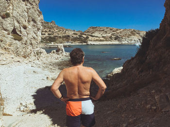 Rear view of shirtless man standing on rock by sea against clear sky